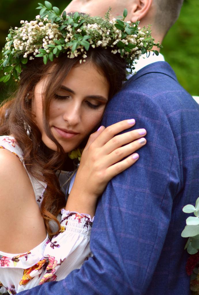 Panna młoda z wiankiem na głowie, z zamkniętymi oczami, przytula się do pana młodego w niebieskim garniturze w kratę.