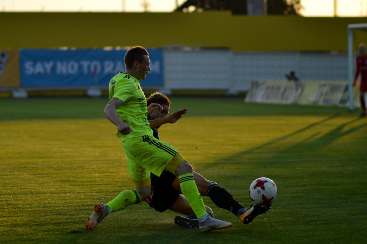 Piłkarz w jaskrawozielonym stroju uderza piłkę stopą, drugi zawodnik próbuje go powstrzymać, w tle żółta trybuna i baner z napisem 'Say No To Racism'.
