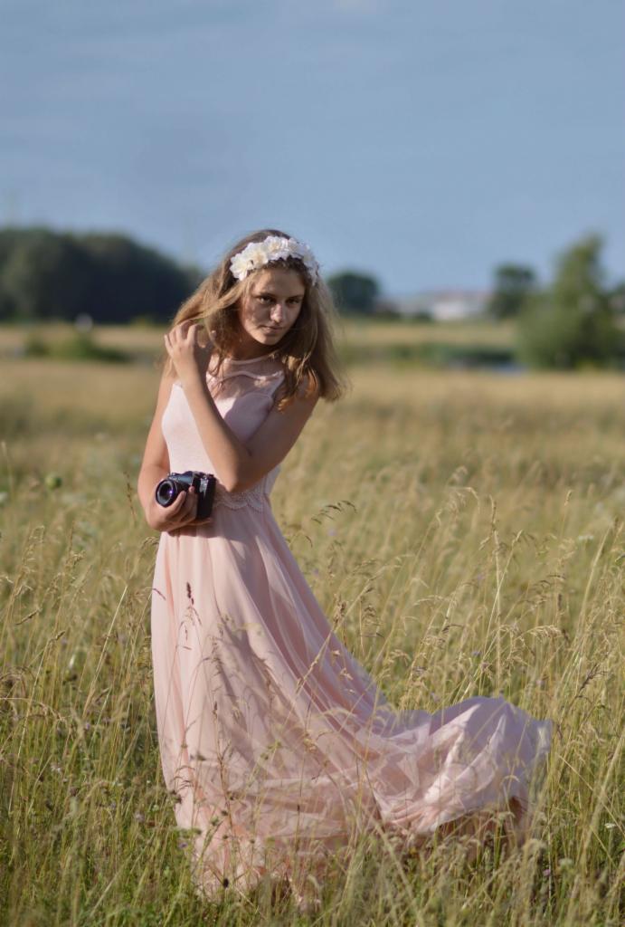Młoda kobieta w zwiewnej, pudrowej sukience i wianku z białych kwiatów, trzymająca stary aparat fotograficzny na tle łąki z wysoką trawą i błękitnego nieba.