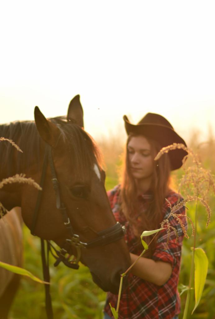 Młoda kobieta w kapeluszu i kraciastej koszuli karmi konia w polu kukurydzy w ciepłym, zachodzącym słońcu.