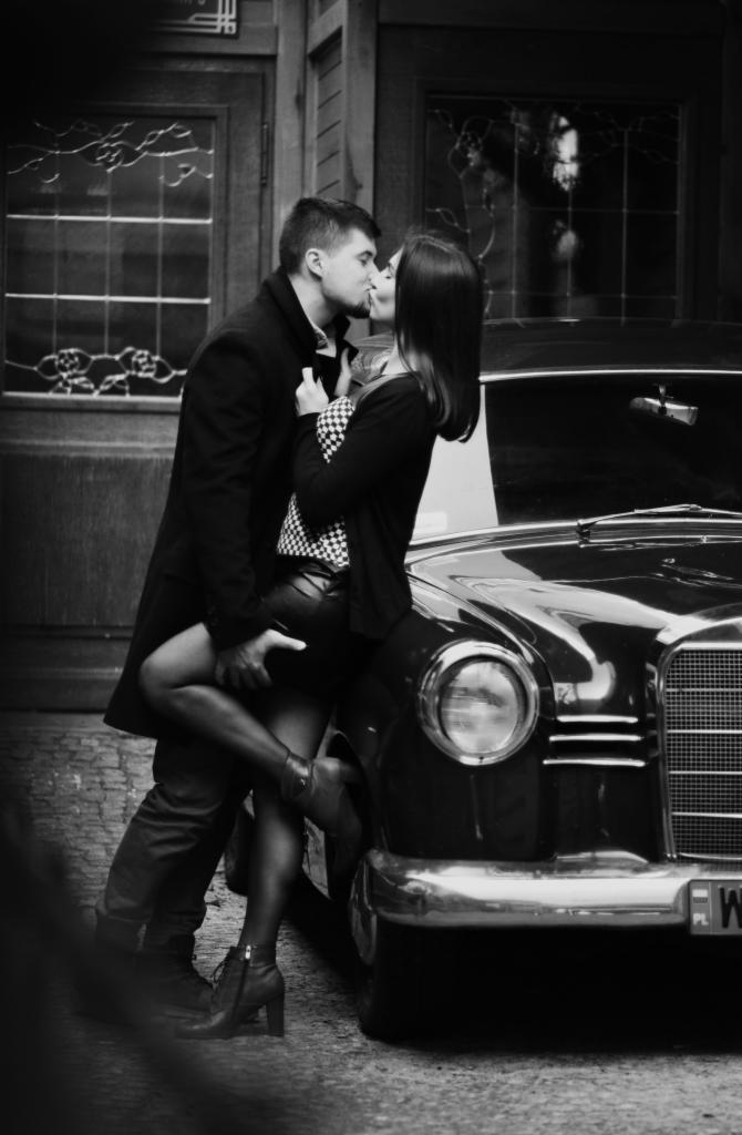 Czarno-białe zdjęcie całującej się pary przy klasycznym samochodzie Mercedes, kobieta w czarnej spódnicy i botkach opiera się o maskę, mężczyzna w płaszczu podtrzymuje ją w talii, w tle drzwi...