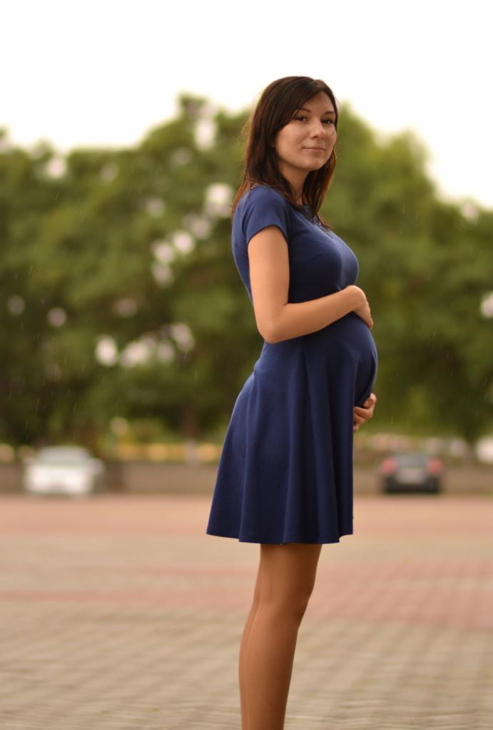 Kobieta w niebieskiej sukience ciążowej, profil, dłonie na brzuchu, zamazane tło miejskie z zielenią i padającym deszczem.