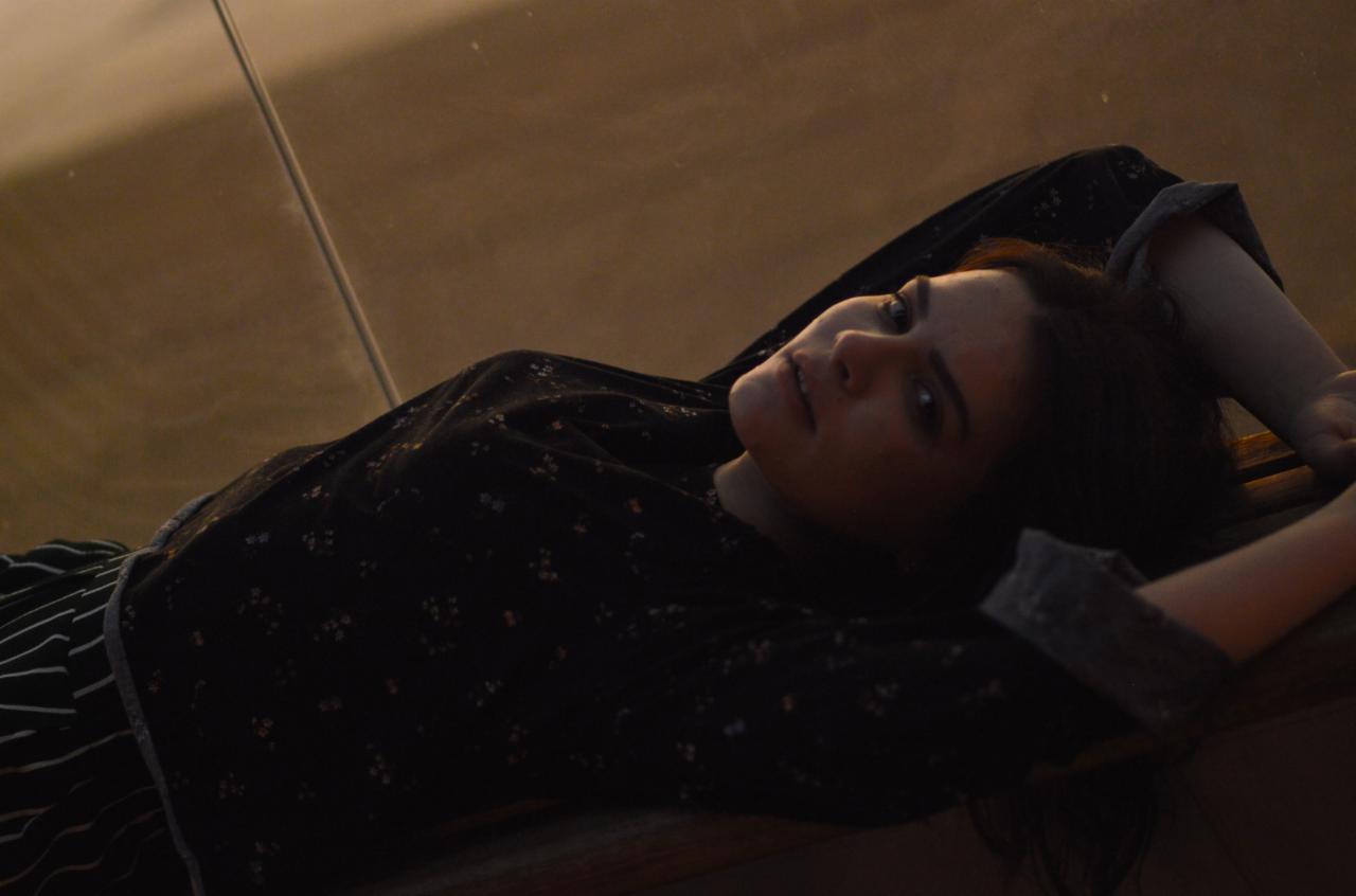 Portret kobiety w ciemnym swetrze z drobnymi kwiatkami, leżącej na drewnianej powierzchni, z rękami założonymi za głową, uchwycony w ciepłym, stonowanym oświetleniu.