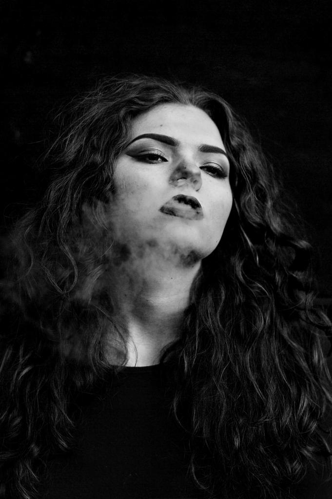 Czarno-białe portretowe ujęcie kobiety z ciemnymi, kręconymi włosami, wydychającej dym z ust, z mocnym makijażem oczu i ust, na ciemnym tle.