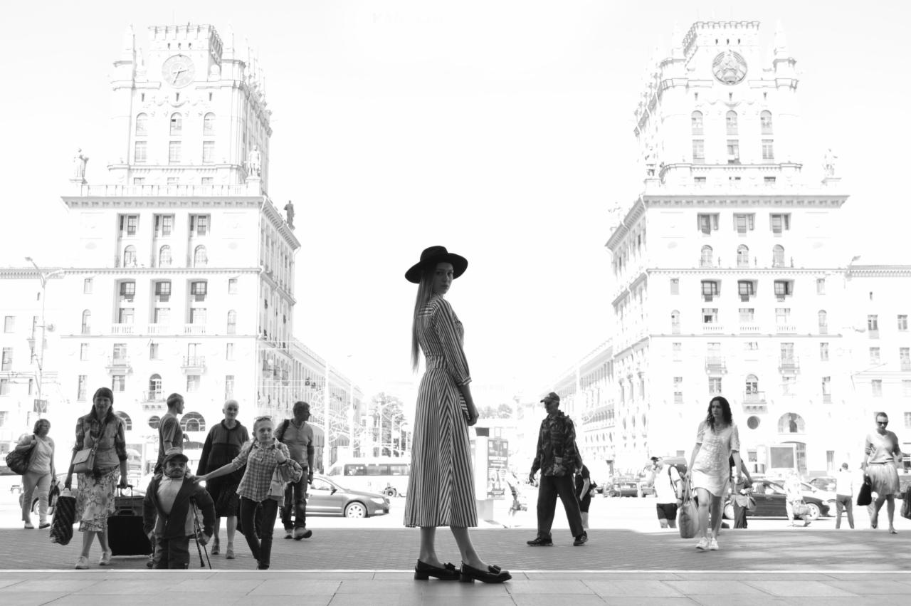 Czarno-białe zdjęcie kobiety w sukience w paski i kapeluszu, stojącej na tle monumentalnych budynków w stylu socrealistycznym, otoczonej przez przechodniów.