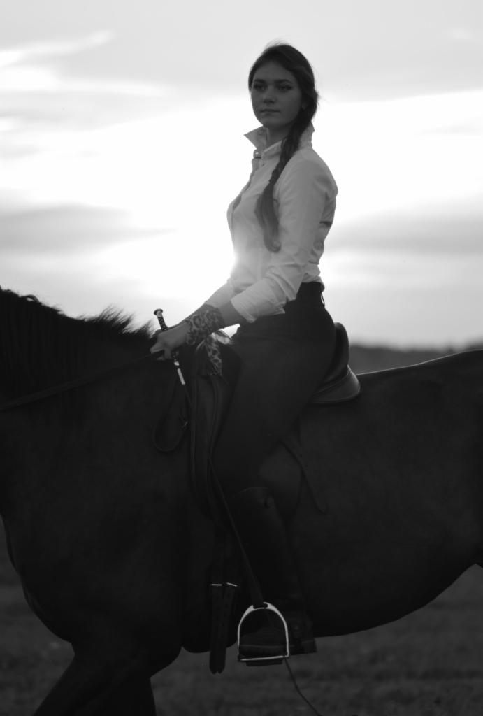 Czarno-białe zdjęcie kobiety z warkoczem i tatuażem na ramieniu, siedzącej na koniu na tle zachodzącego słońca.