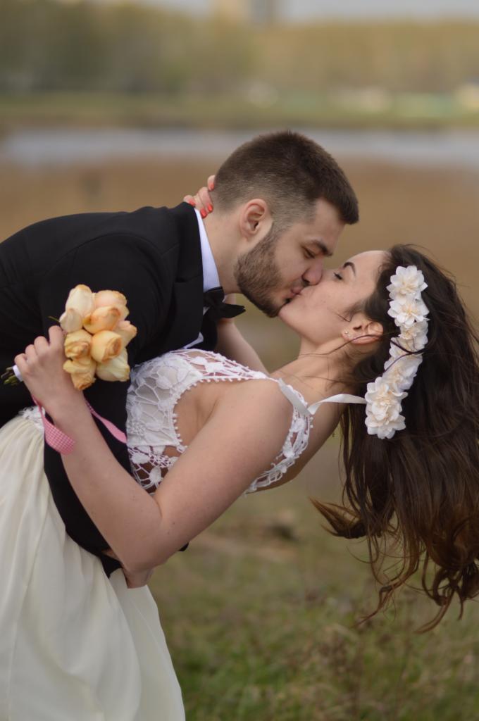 Pocałunek młodej pary: panna młoda w koronkowej sukni i wianku z białych kwiatów, trzyma bukiet brzoskwiniowych róż, pochylona w ramionach pana młodego w czarnym garniturze.