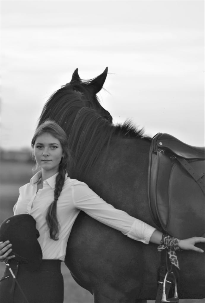 Portret młodej kobiety w białej koszuli i czarnych spodniach, trzymającej kask jeździecki, opierającej się o ciemnego konia z siodłem; zdjęcie w czerni i bieli.