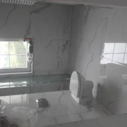 Wykończona łazienka z białymi płytkami imitującymi marmur, widoczna toaleta, okno z widokiem na zieleń i częściowo zasłonięta kabina prysznicowa.