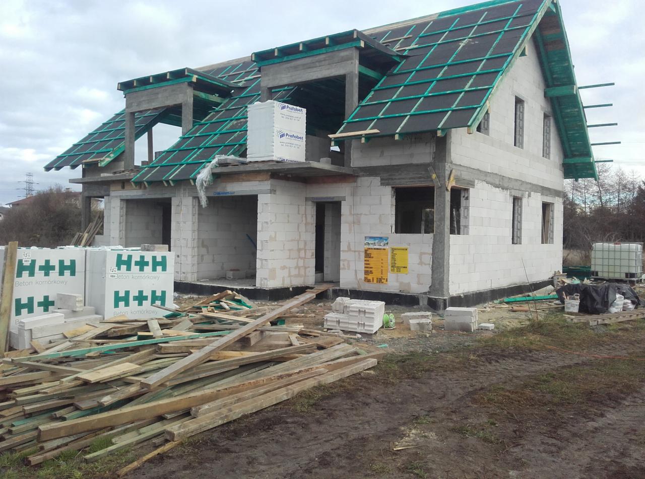 Surowy stan dwukondygnacyjnego domu z betonu komórkowego H+H, z widoczną konstrukcją dachu pokrytą czarną papą i zielonymi łatami, palety z materiałem budowlanym na placu budowy, pochmurne niebo...