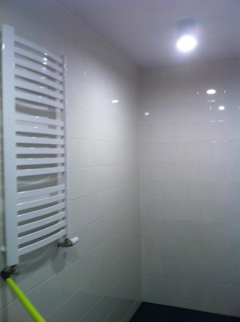 Biały, drabinkowy grzejnik łazienkowy na ścianie wyłożonej jasnymi, błyszczącymi płytkami w małej łazience z czarną podłogą.