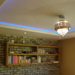 Nowoczesne wnętrze z ceglaną ścianą, drewnianą półką z książkami i ozdobnym żyrandolem podświetlonym niebieskim światłem LED w suficie.