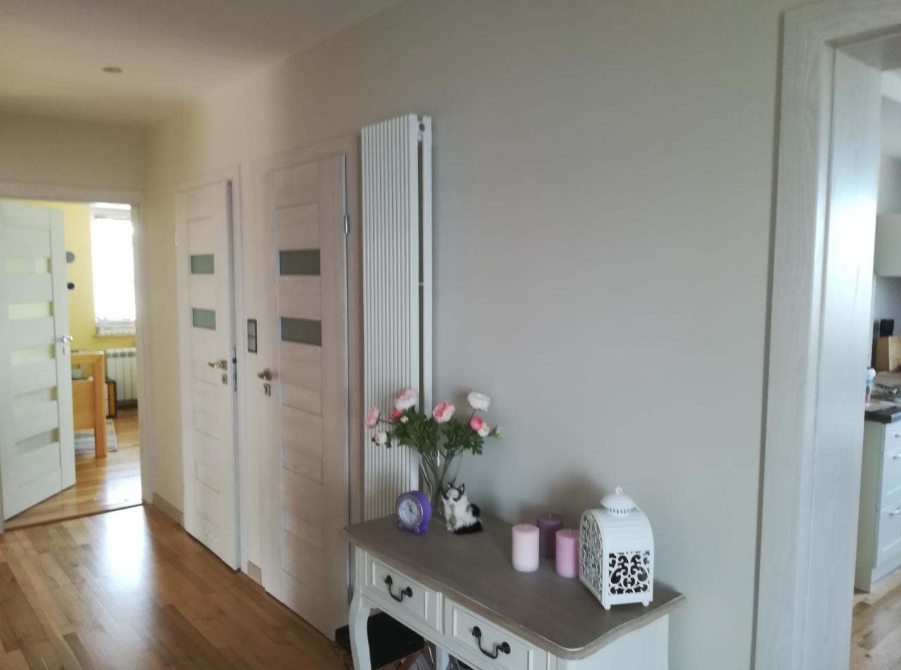 Jasnoszary korytarz z drewnianą podłogą, białymi drzwiami z przeszkleniami i grzejnikiem panelowym. Dekoracyjny stolik z kwiatami, zegarem i lampionem pod ścianą.