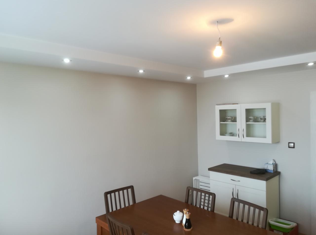 Jasne, świeżo pomalowane wnętrze jadalni z drewnianym stołem, krzesłami, i wiszącą szafką z porcelaną, oświetlone punktowymi lampami LED i pojedynczą żarówką.