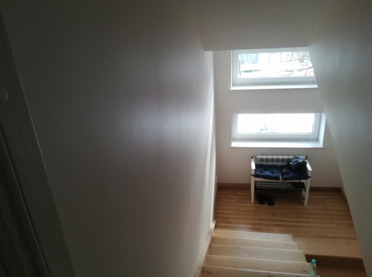 Świeżo pomalowana, jasnoszara ściana klatki schodowej z drewnianymi schodami, widoczne dwa okna dachowe i biały regał na buty z poduszkami i torbą.