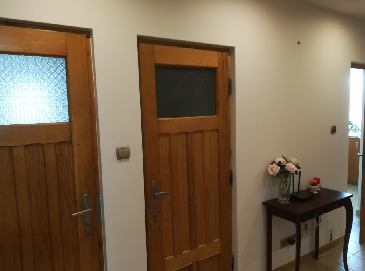 Dwie drewniane drzwi wewnętrzne z szybami, pomalowane ściany na biało, na stoliku wazon z różowymi kwiatami i router.