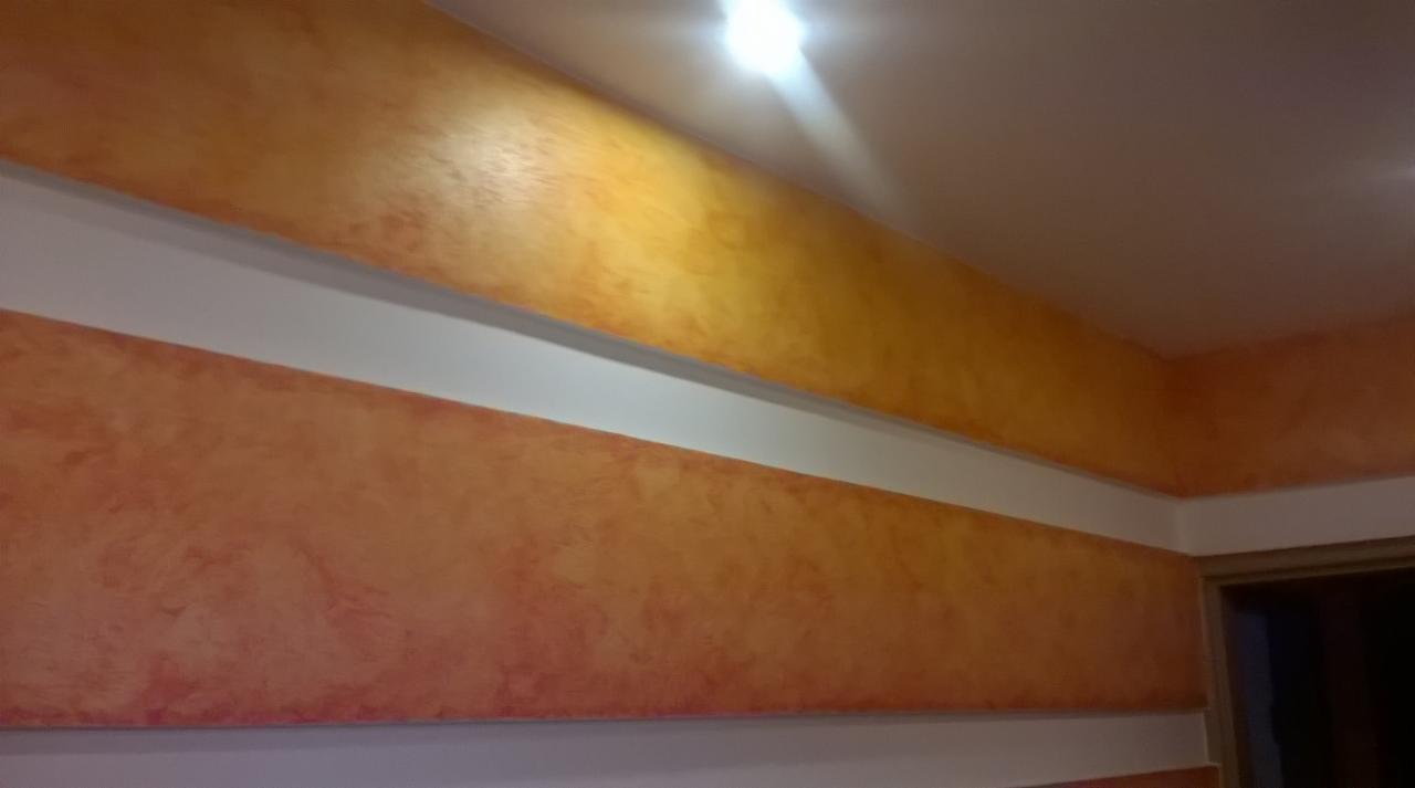 Pomarańczowa ściana z dekoracyjną strukturą tynku, oddzielona białymi pasami, oświetlona górnym światłem.