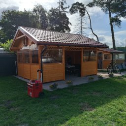 WORK IN WOOD Grzegorz Rachubiński - Drewniana konstrukcja z brązowym dachem i przezroczystymi ścianami, prawdopodobnie altana lub zadaszone miejsce do siedzenia, z widocznym wyposażeniem wewnątrz, skrzynką narzędziową na zewnątrz...