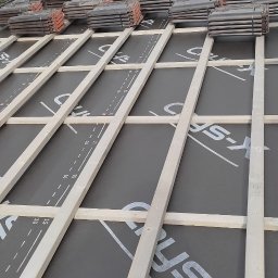 WORK IN WOOD Grzegorz Rachubiński - Etap budowy dachu: widok na konstrukcję drewnianą z membraną dachową z nadrukowaną miarką, przygotowaną pod dachówki, z widocznym stosem dachówek w tle i fragmentem orynnowania.