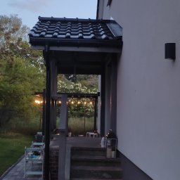 WORK IN WOOD Grzegorz Rachubiński - Zadaszone wejście do domu z czarnym, ceramicznym dachem, widok na oświetlony sznurem żarówek taras i ogród z huśtawką w tle, wczesny wieczór.