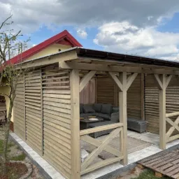 Drewniana altana z lamelami w Radowie Małym, wyposażona w szarą sofę i stolik kawowy, z widocznym fragmentem ogrodu i budynku z czerwonym dachem w tle.