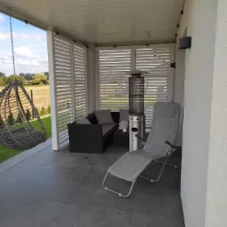 Zadaszony taras w Radowo Małe z meblami ogrodowymi, wiszącym fotelem i promiennikiem ciepła, osłonięty żaluzjami.