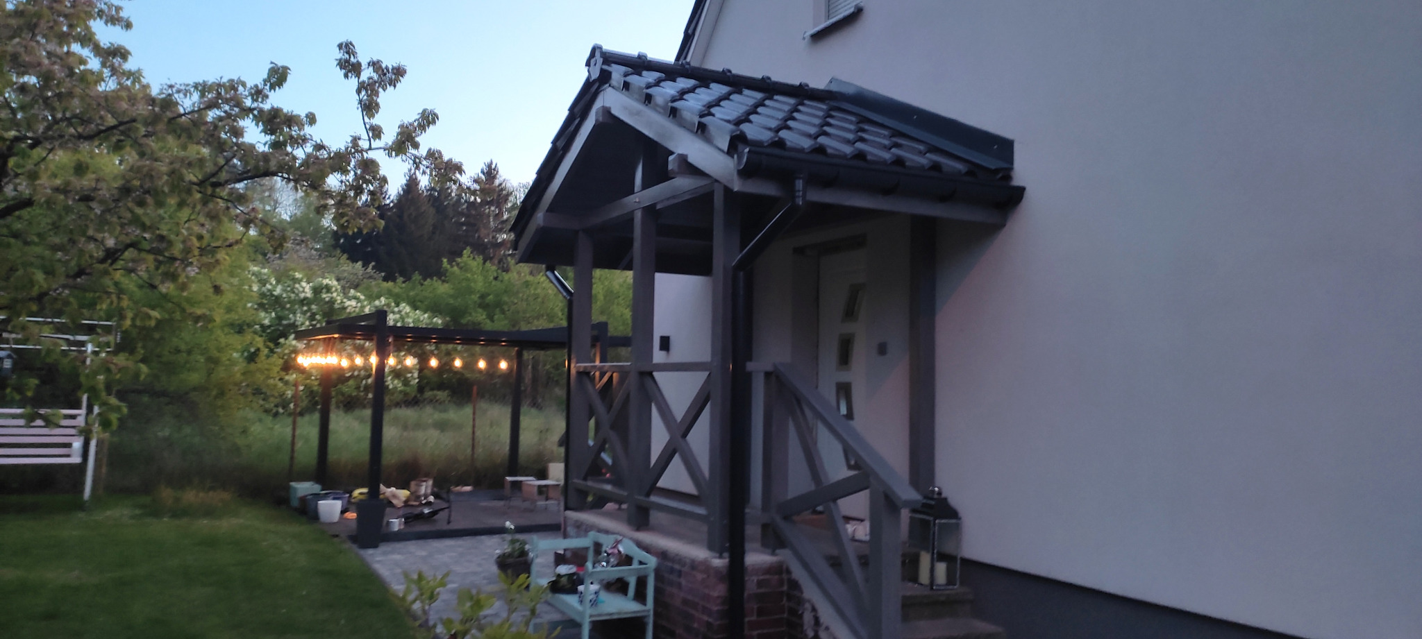 Zadaszone wejście do domu z drewnianą konstrukcją, widoczna pergola z oświetleniem w tle, wczesny wieczór.