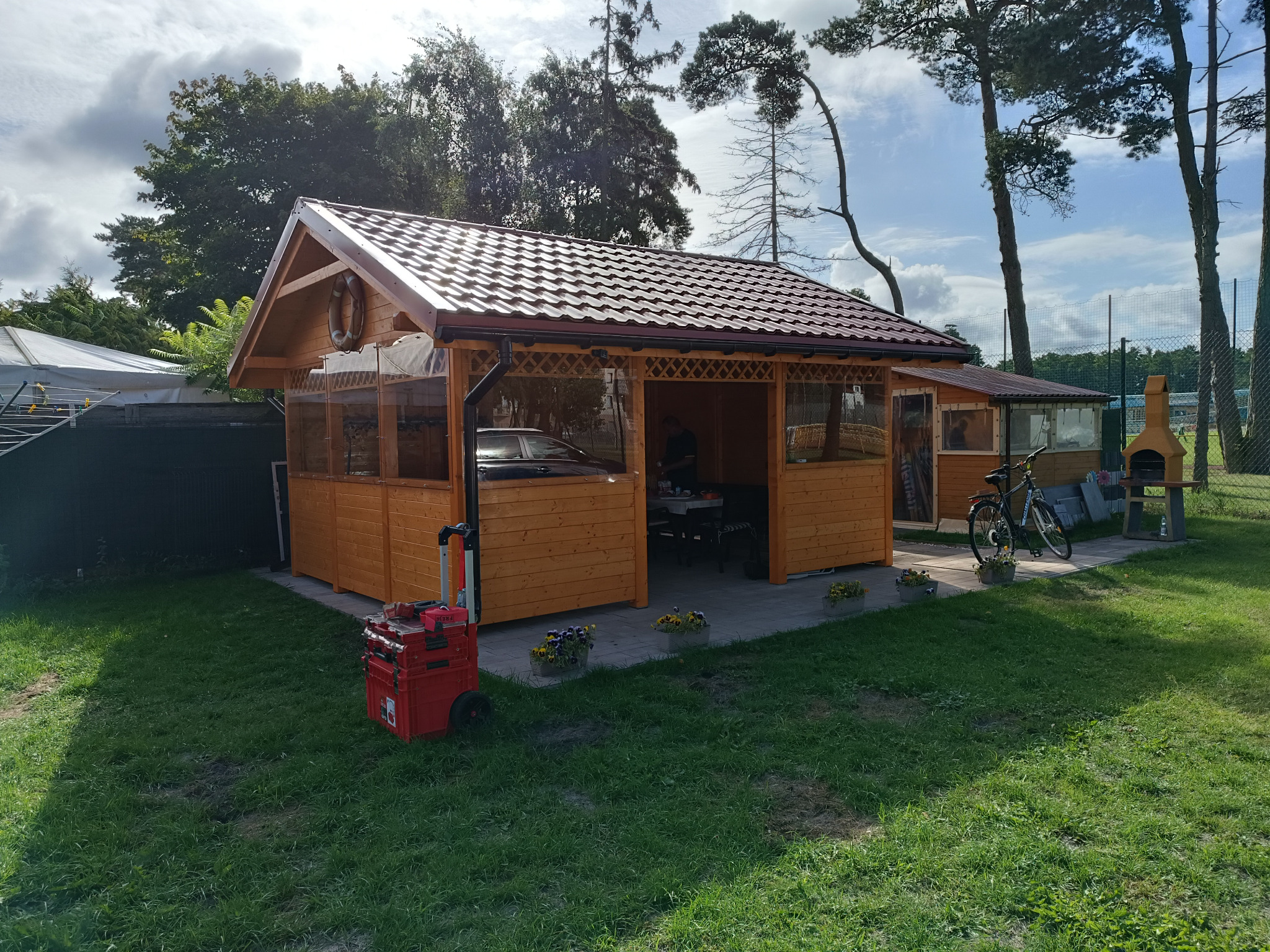 Drewniana konstrukcja z brązowym dachem i przezroczystymi ścianami, prawdopodobnie altana lub zadaszone miejsce do siedzenia, z widocznym wyposażeniem wewnątrz, skrzynką narzędziową na zewnątrz...