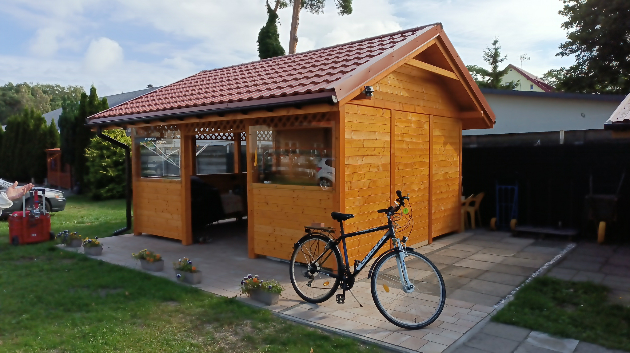 Drewniana altana z czerwonym dachem, częściowo przeszklona, z rowerem zaparkowanym przed nią, otoczona kostką brukową i doniczkami z kwiatami.