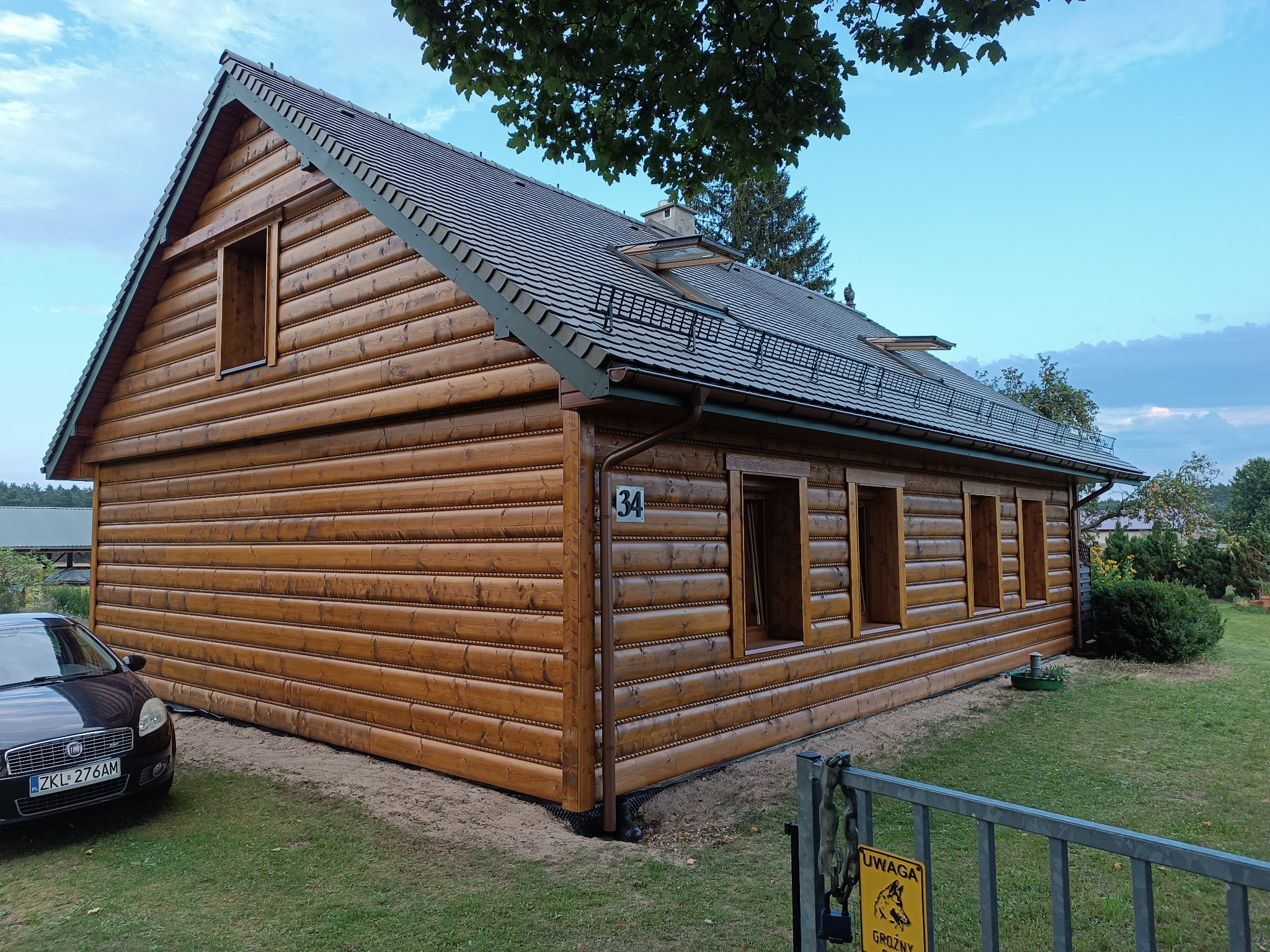 Drewniany dom w stylu góralskim z ciemnobrązową elewacją z bali, szary dach z lukarnami, numer domu 34, widoczny samochód osobowy i fragment ogrodzenia z tabliczką ostrzegawczą