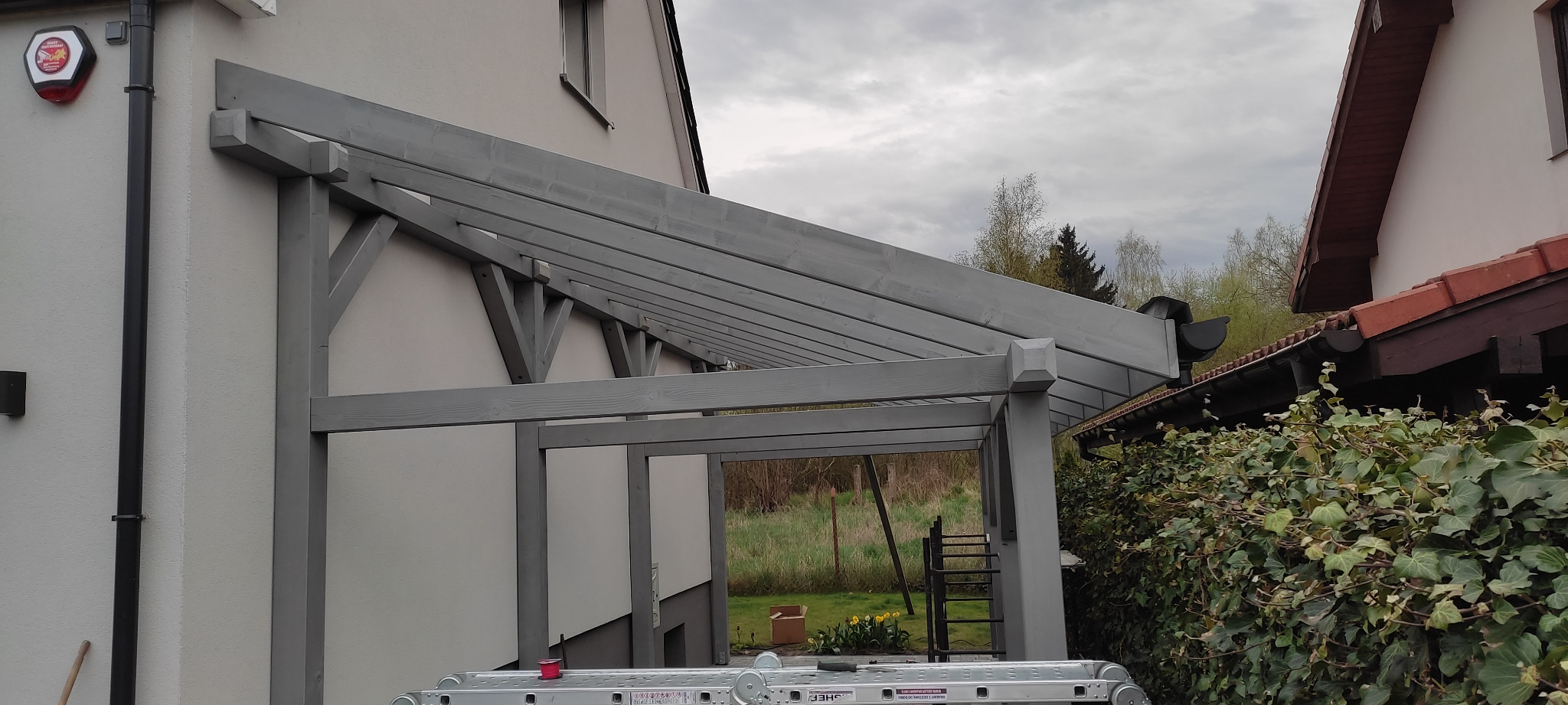 Szara pergola przylegająca do ściany domu, widok z dołu, drabina oparta o konstrukcję, w tle zieleń ogrodu i fragment dachu sąsiedniego budynku.