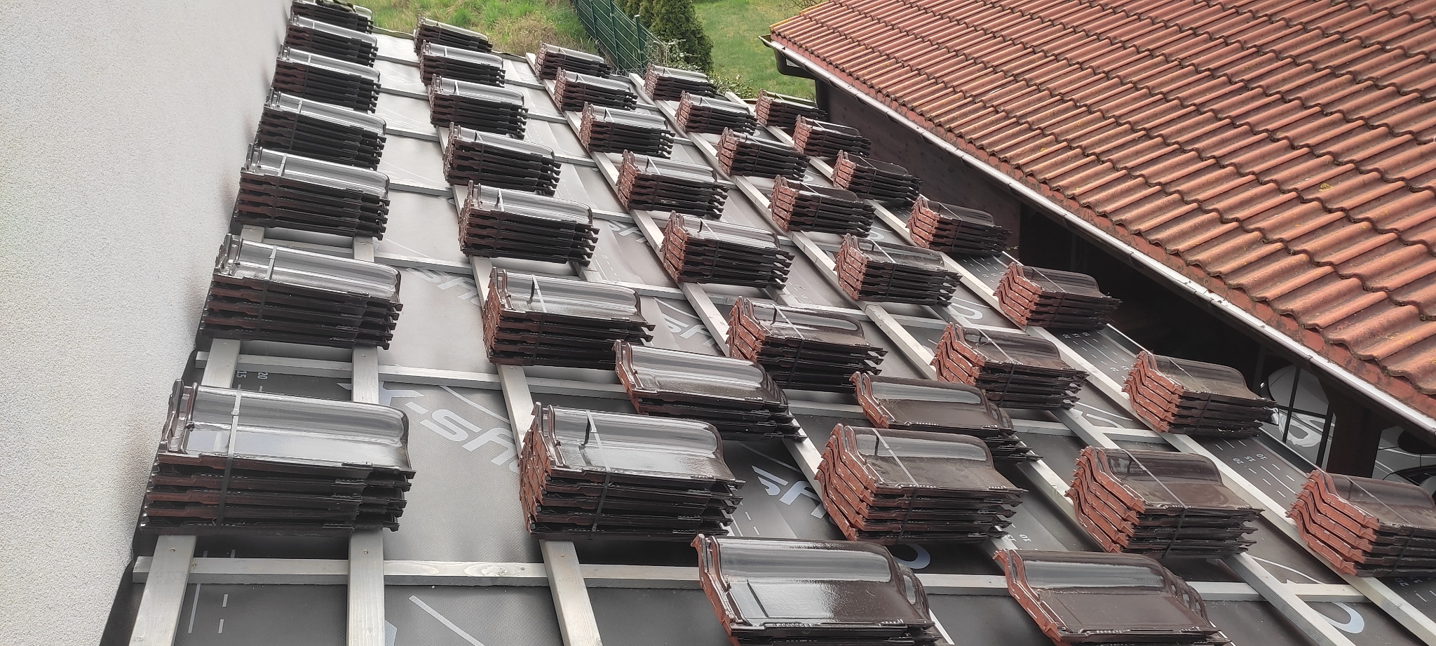 Przygotowane pakiety ceramicznej dachówki karpiówki ułożone rzędami na łatach i kontrłatach, gotowe do krycia dachu, widoczny fragment istniejącego dachu z dachówki ceramicznej.