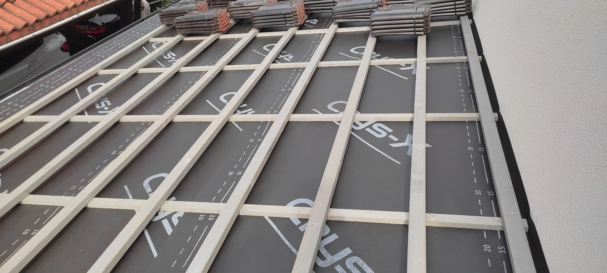 Etap budowy dachu: widok na konstrukcję drewnianą z membraną dachową z nadrukowaną miarką, przygotowaną pod dachówki, z widocznym stosem dachówek w tle i fragmentem orynnowania.