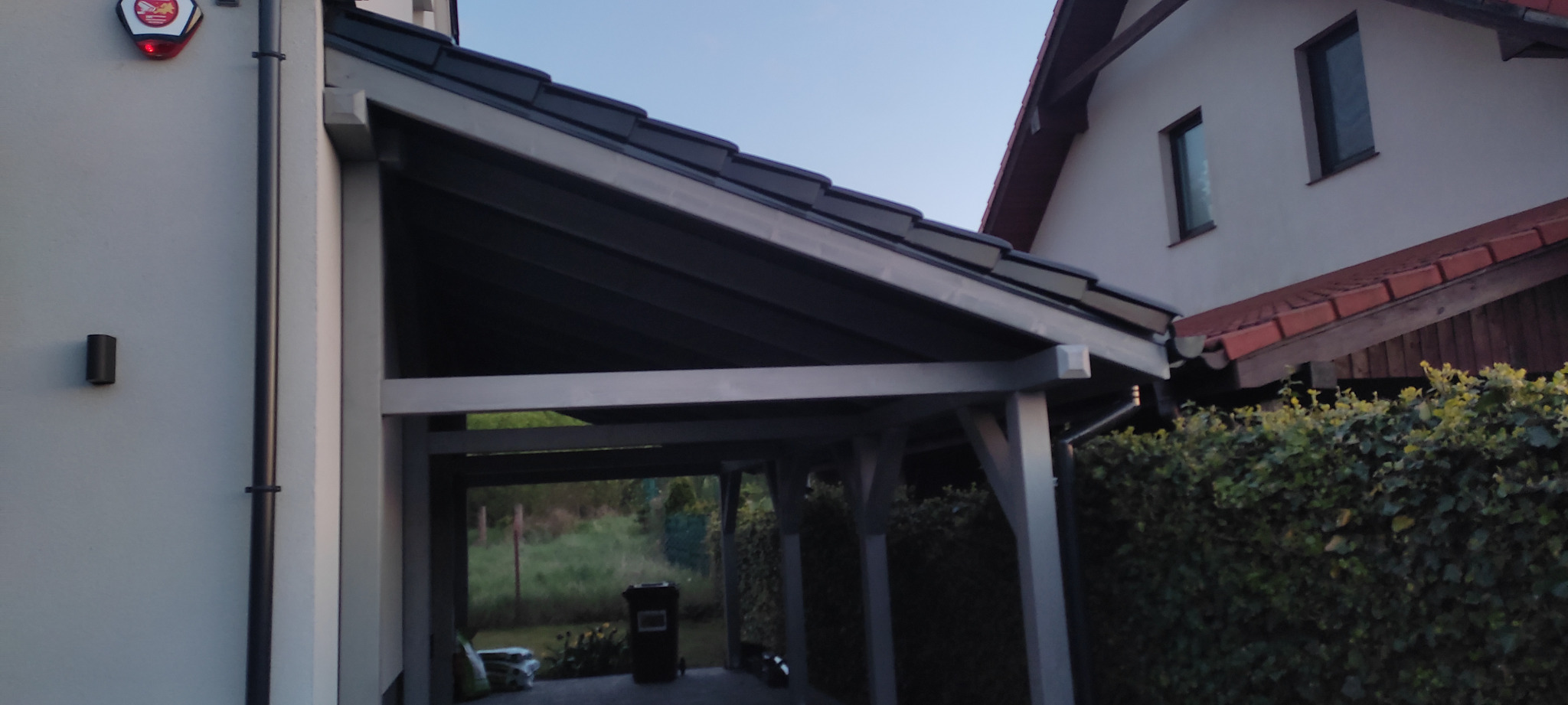 Drewniana konstrukcja z zadaszeniem w kolorze grafitowym, przylegająca do budynku z alarmem, widok na ogród w tle.