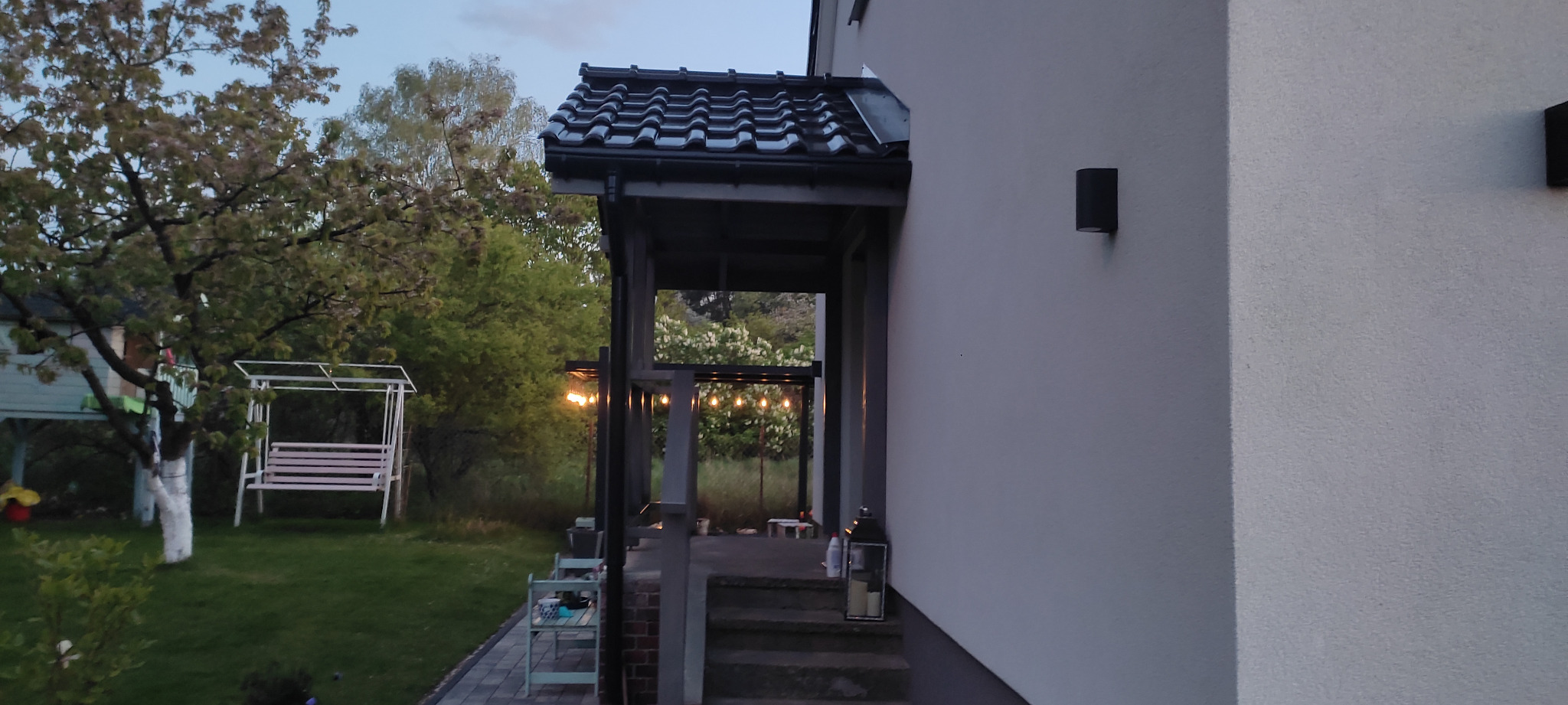 Zadaszone wejście do domu z czarnym, ceramicznym dachem, widok na oświetlony sznurem żarówek taras i ogród z huśtawką w tle, wczesny wieczór.