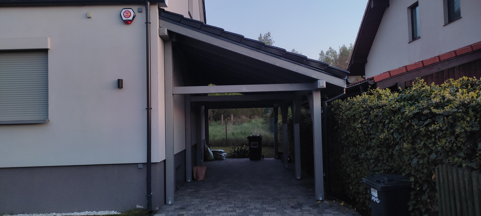 Zadaszone miejsce parkingowe przy domu z kostki brukowej, widoczne worki z ziemią w tle i alarm na ścianie budynku.