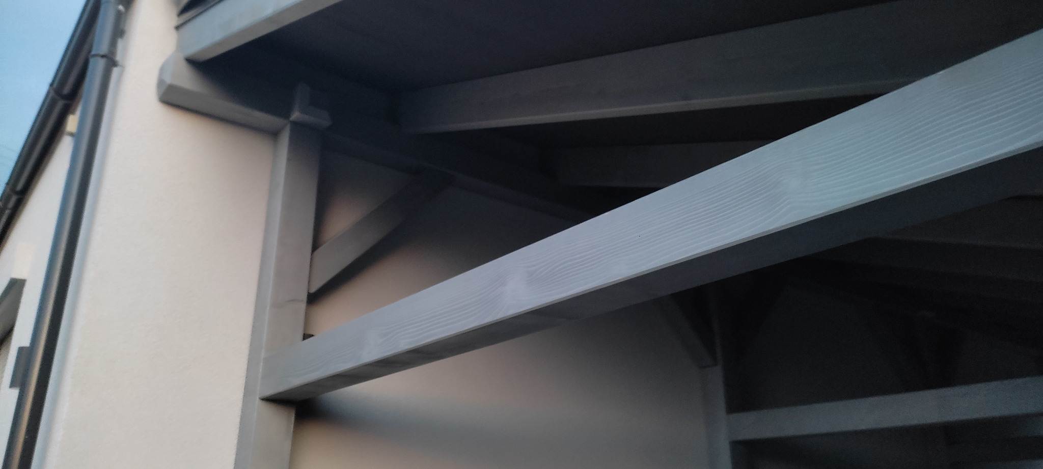 Fragment szarej drewnianej konstrukcji tarasu przylegającego do ściany budynku z widoczną rynną, ujęcie od dołu.