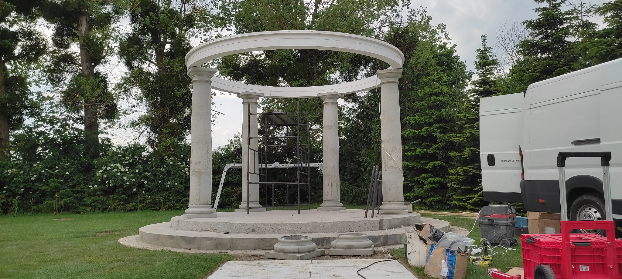 Niedokończona altana z kolumnami i okrągłym dachem na betonowej podstawie, widoczne rusztowanie, narzędzia, biały bus w tle i zieleń ogrodu.