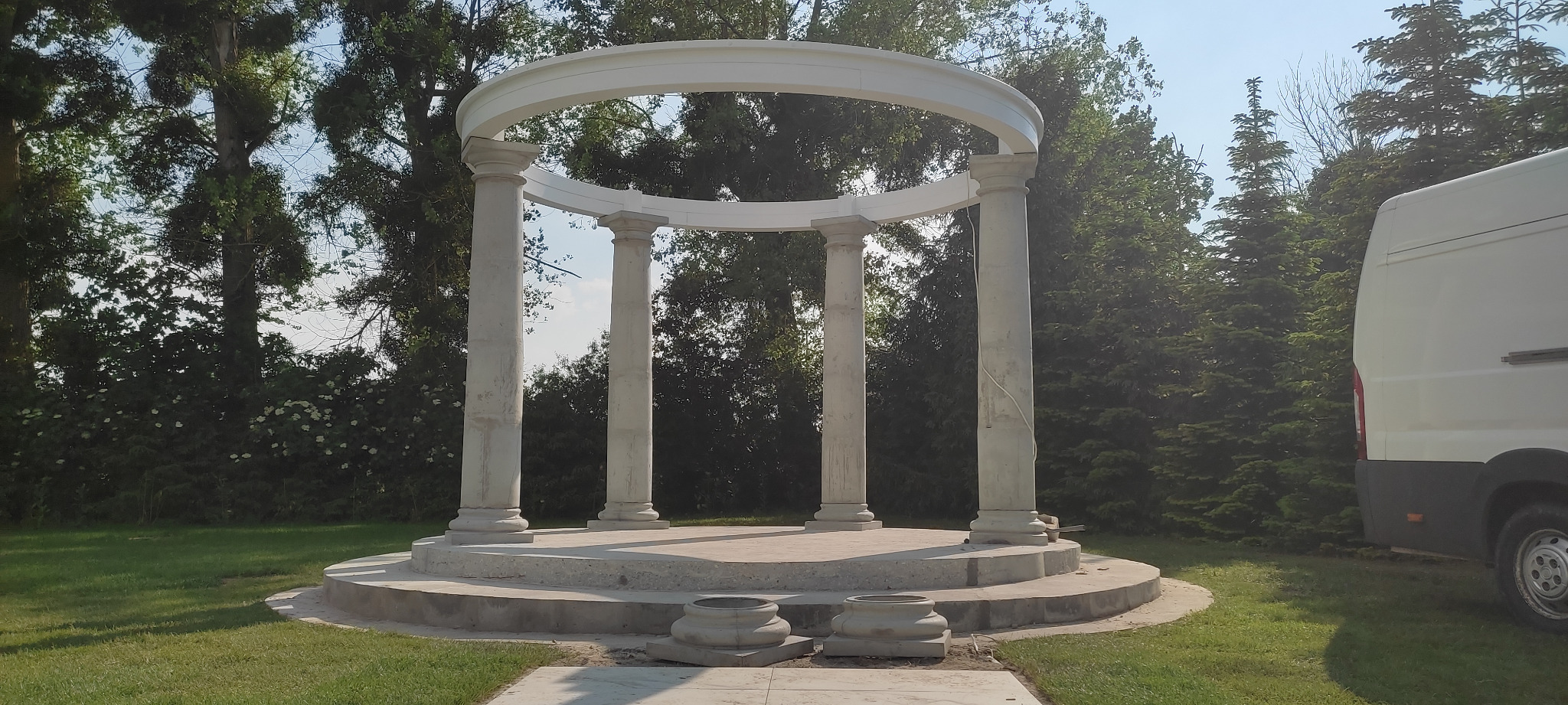 Biała, okrągła altana z kolumnami w ogrodzie, widoczny fragment białego samochodu dostawczego po prawej stronie.