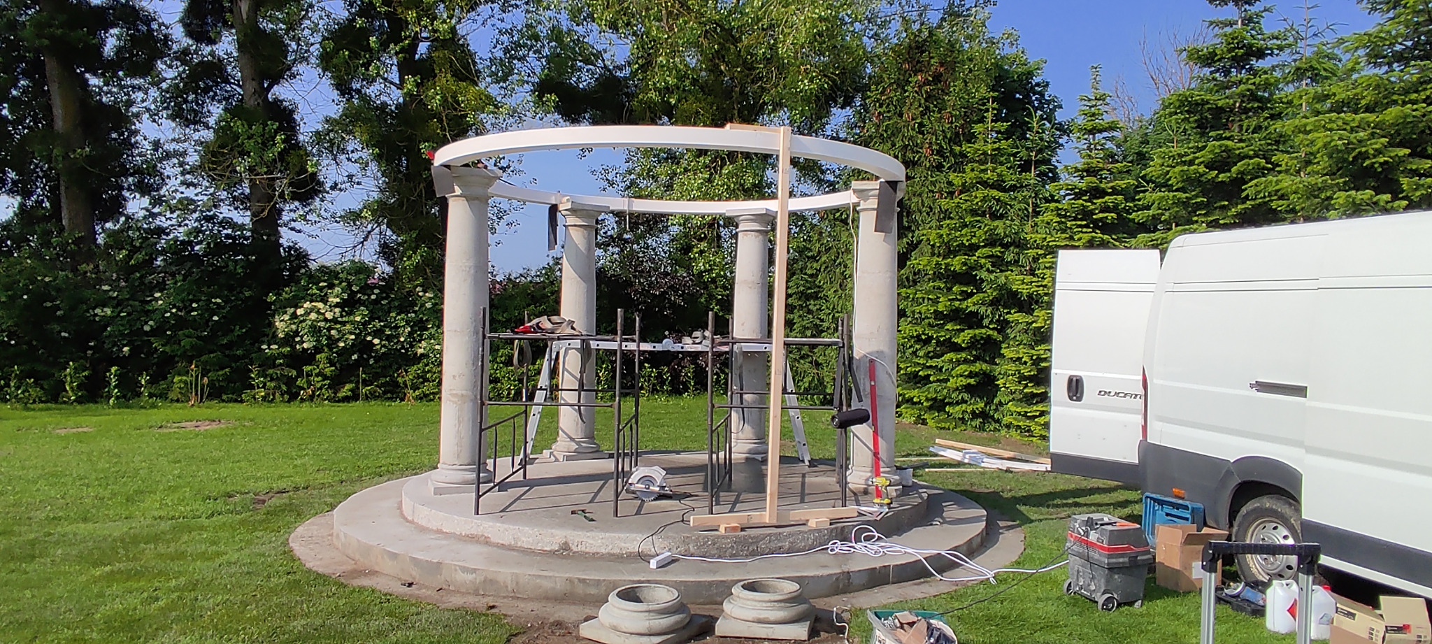 Budowa altany ogrodowej z kolumnami: widoczne rusztowania, narzędzia i podstawa konstrukcji na tle zielonego trawnika i drzew, częściowo zasłonięta przez biały samochód dostawczy.