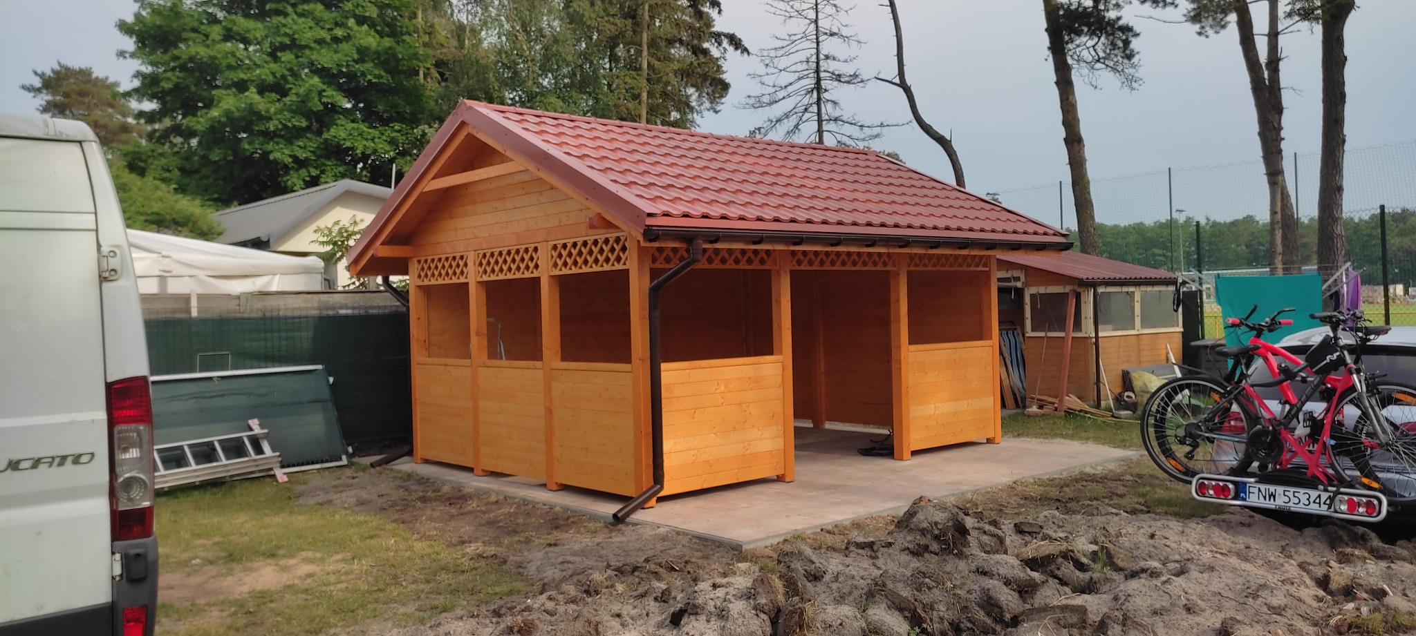 Świeżo postawiona drewniana altana z czerwoną dachówką, widoczna z boku, z samochodem dostawczym i rowerami w tle, teren wokół w trakcie zagospodarowywania.