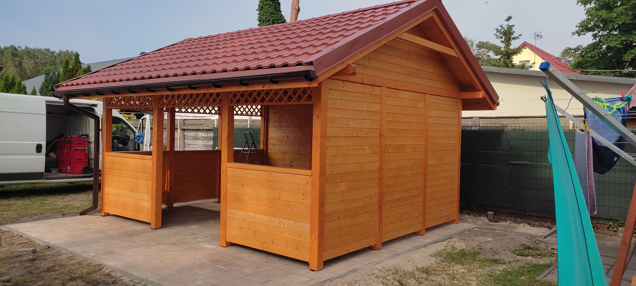 Drewniana altana z czerwonym dachem na kostce brukowej, widoczny samochód dostawczy z narzędziami i drabina wewnątrz konstrukcji.