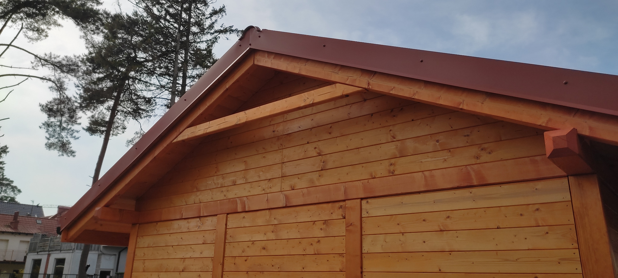 Fragment drewnianej konstrukcji z brązowym dachem, widok z dołu, na tle drzew i zabudowań.