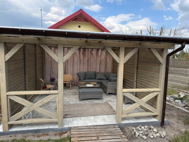 Zadaszona konstrukcja ogrodowa z drewnianą ramą, wyposażona w szarą sofę narożną, stolik kawowy z rattanu, drewniane krzesła i zasłony, posadowiona na kostce brukowej, z widocznym fragmentem domu...