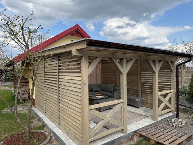 Drewniana altana z lamelami w Radowie Małym, wyposażona w szarą sofę i stolik kawowy, z widocznym fragmentem ogrodu i budynku z czerwonym dachem w tle.