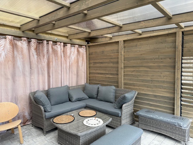 Zadaszona przestrzeń wypoczynkowa z drewnianą konstrukcją, zasłonięta zasłonami w motywy roślinne, wyposażona w szary rattanowy narożnik, stolik kawowy i krzesło z jasnego drewna.