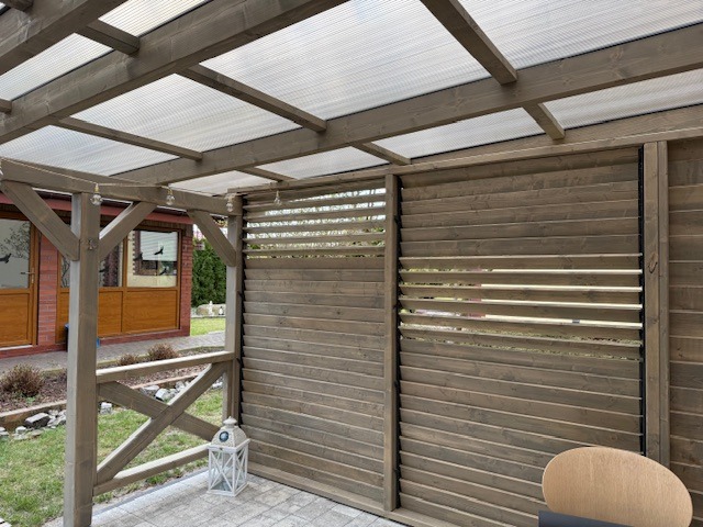 Zadaszona pergola drewniana z ażurową ścianą boczną, oświetlona girlandą żarówek, z widocznym fragmentem domu w tle.