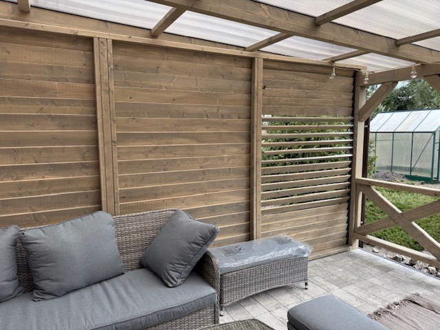 Zadaszona konstrukcja z drewnianymi panelami i ażurowymi przesłonami, szara sofa ogrodowa z poduszkami, widoczny fragment tarasu z kostki brukowej oraz szklarnia w tle.