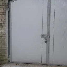 Szara, dwuskrzydłowa brama garażowa z zamkiem na tle ceglanego muru z łukowatym nadprożem.