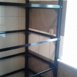 Czarna, metalowa konstrukcja regału z półkami, zamontowana w rogu pomieszczenia z wyłożonymi płytkami ścianami, widoczne śruby mocujące.